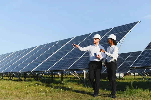 Sert şapka ve üniforma giyen mühendisler güneş panellerini inceliyor, temiz enerji ve sürdürülebilir teknoloji üzerinde işbirliği yapıyorlar.