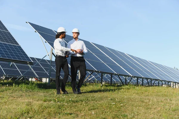 Güneşli bir günde güneş panellerinin önünde durarak yenilenebilir enerji projesini tartışan iki farklı mühendis.
