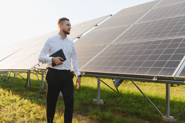 Gömlekli ve pantolonlu bir adam güneş panellerinden geçiyor, elinde dijital bir tablet, sürdürülebilir bir enerji projesini yönetiyor.