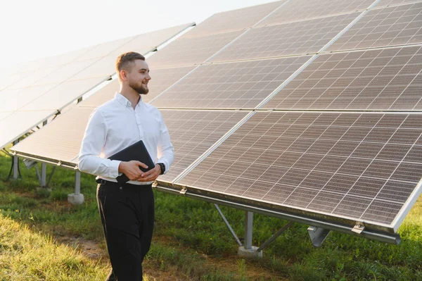 Beyaz gömlekli ve siyah pantolonlu bir adam elinde tabletle yenilenebilir enerji santralinin güneş panellerinden geçiyor.