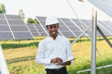 Dijital tablet tutan ve güneş paneli alanında gülümseyen, kasklı Afrikalı Amerikalı bir adam. Yenilenebilir enerji kavramı