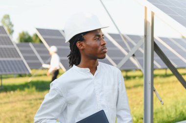 Başlık takan genç mühendis, elinde defter, güneş enerjisi istasyonundaki yenilenebilir enerji tesisatını denetliyor.