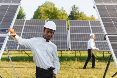 Genç siyah adam kasketli ve beyaz gömlekli fotovoltaik panelleri kontrol ediyor, arka planda yürüyen bir meslektaşı var.