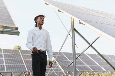 Sert bir şapka takan Afrikalı mühendis büyük bir yenilenebilir enerji alanındaki güneş panellerini geziyor ve denetliyor.