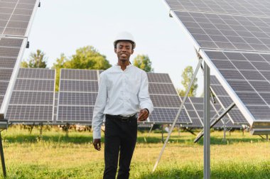 Siyah şapkalı Afrikalı Amerikalı mühendis, güneş panelleri arasında yürüyor ve gülümsüyor, yenilenebilir enerji üretiyor.