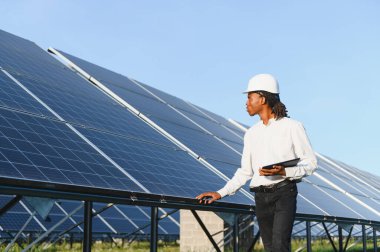 Sert bir şapka takan ve elinde tablet tutan genç bir adam gün boyunca bir elektrik santralinde güneş panellerini kontrol ediyor.