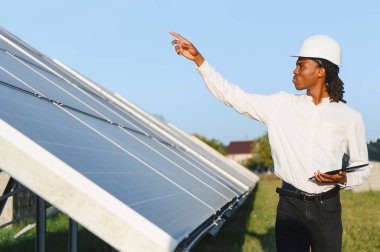 Sert bir şapka takan ve elinde tablet olan Afrikalı mühendis yenilenebilir enerji üretimi için güneş paneli alanını inceliyor.