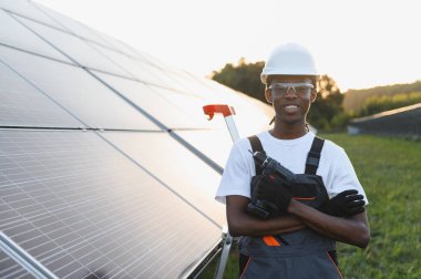 Güvenlik ekipmanı ve üniforma giyen, elinde matkap tutan, güneş panellerinin önünde duran Afrikalı Amerikalı teknisyen.