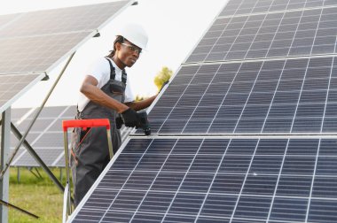 Siyah şapka ve güvenlik gözlüğü takan Afrikalı Amerikalı teknisyen yenilenebilir enerji için güneş panelleri kuruyor.