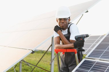 Siyah şapkalı ve güvenlik gözlüklü Afro-Amerikan adam elinde elektrikli matkap, güneş paneli kurulumu üzerinde çalışıyor.