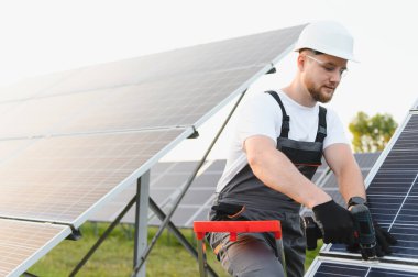 Sert bir şapka ve güvenlik gözlüğü takan erkek işçi dışarıda matkapla bir güneş paneli kuruyor ve yeşil enerji üretiyor.