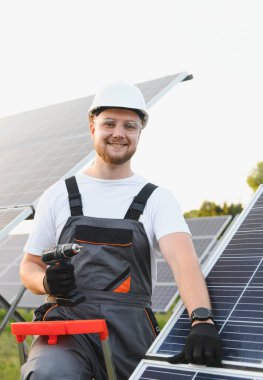 Genç teknisyen güneş çiftliğine fotovoltaik paneller kuruyor, yenilenebilir enerji üretimi için elektrikli matkapla çalışıyor.