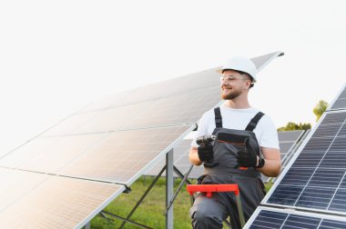 Elinde matkap tutan kendinden emin teknisyen güneş enerjisi çiftliğine fotovoltaik paneller yerleştiriyor ve yenilenebilir enerji gelişimini temsil ediyor.