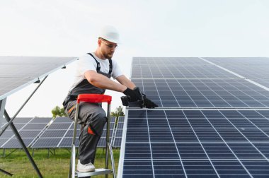 Tulumlu ve kasklı bir adam güneş çiftliğinde çalışıyor, yeşil enerji üretimi için fotovoltaik paneller kuruyor.