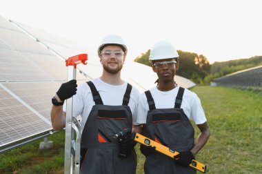Kurulum sırasında güvenlik ekipmanı giyen iki farklı mühendis güneş paneli alanının önünde duruyor ve ellerinde aletler var.