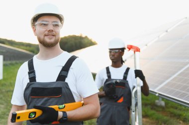 Güneş enerjisi santralinde çalışan ve fotovoltaik paneller kuran iki farklı kasklı ve güvenlik gözlüklü teknisyen.