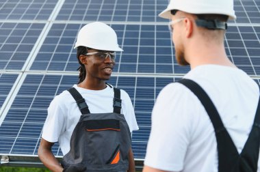 İki mühendis, güneş enerjisi santralinde, panellerin önünde çalışmaktan bahsediyorlar.