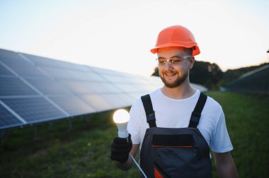 Elinde parlak bir ampul tutan mühendis fotovoltaik panellerin önünde duruyor, yenilenebilir enerji ve yeniliği simgeliyor.