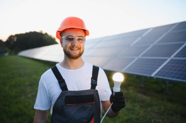 Elinde parlayan bir ampul tutan gülen mühendis, yeşil bir alanda güneş panellerinin ürettiği temiz enerjiyi sembolize ediyor.