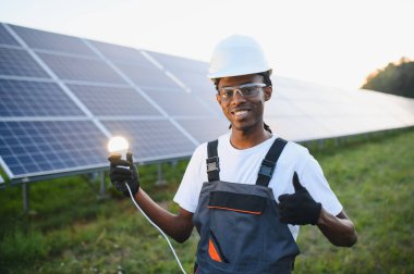Afrikalı Amerikalı teknisyen gülümsüyor, elinde ampulü tutuyor, güneş panellerinin önünde başparmağını kaldırıyor.