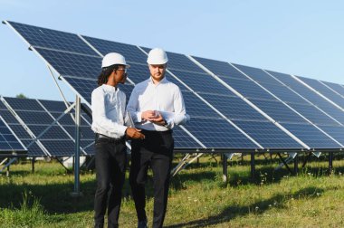 Güneş panellerini teftiş eden, açık mavi gökyüzünün altındaki yenilenebilir enerji tesisinde çalışan mühendisler.