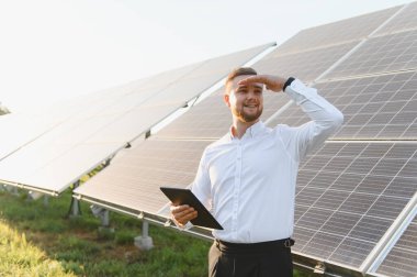 Genç mühendis bir tablet tutuyor, gözleri koruyor, güneş panellerinin işleyişini denetliyor, yeşil enerji üretimine katkıda bulunuyor.