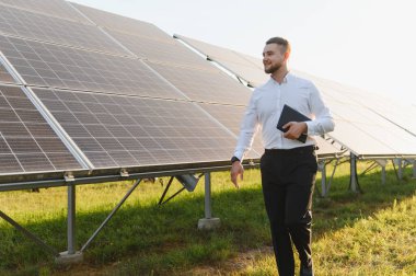 Gömlekli ve pantolonlu bir adam temiz güneş panellerinin arasında tabletle yürüyor, sürdürülebilir güç kurulumunu inceliyor.