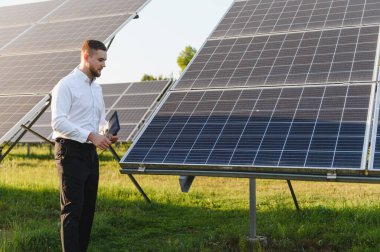 Genç adam yeşil alanda bir tablet kullanarak güneş paneli performansını izliyor, yenilenebilir enerji ve sürdürülebilirliğe odaklanıyor