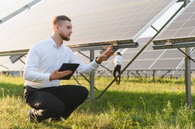 Güneş panellerini kontrol eden mühendis, yenilenebilir enerji verilerini kaydetmek için dijital tablet tutuyor.
