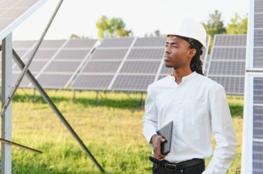 Siyah adam elinde tablet tutan, güneş enerjisi santralinde duran, yenilenebilir enerji konsepti olan...