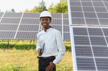 Afrikalı Amerikalı mühendis kendinden emin görünüyor, şapka takıyor, modern bir güneş paneli alanının önünde duruyor.