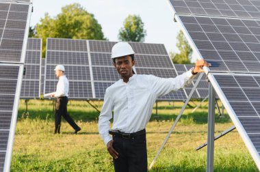 Bir güneş paneli alanında çalışan Afrikalı Amerikalı mühendis yenilenebilir enerji üretimini denetliyor. Arka planda başka bir işçi var.