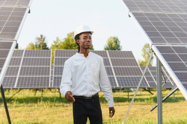 Sert bir şapka takan Afrikalı Amerikalı mühendis güneş panellerini denetliyor yenilenebilir enerji ve sürdürülebilirliği sembolize ediyor.