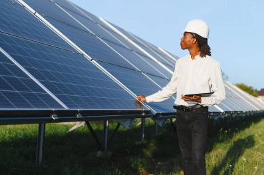 Afrika kökenli Amerikalı mühendis başlık takıyor, elinde tablet tutuyor ve güneşli bir alandaki güneş panellerini inceliyor.