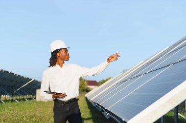 Güneş panellerini denetleyen, elinde tablet tutan ve yenilenebilir enerji çözümlerini analiz eden bir başlık içindeki Afrikalı mühendis.
