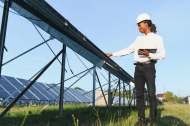 Başlık takan ve elinde tablet tutan genç bir adam yeşil bir alanda güneş panellerini inceliyor.