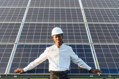 Siyah şapkalı genç adam gülümsüyor, yenilenebilir enerji için büyük güneş panellerinin önünde güvenle duruyor.