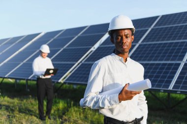 Kasklı genç mühendis elinde planlar, güneş panellerinin önünde duruyor ve meslektaşı verileri kontrol ediyor.