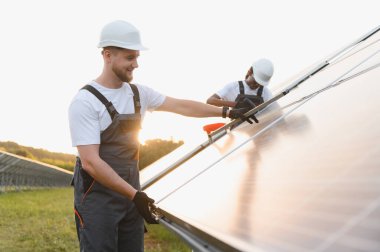 Erkek teknisyenler güneş panellerini kurup temizliyor. Gün batımında bir tarlada yenilenebilir elektrik üretiyorlar.
