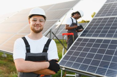 Genç teknisyen gülümserken ekip üyesi güneşli bir günde yenilenebilir enerji santraline güneş panelleri kuruyor.