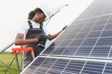 Güçlü şapka ve güvenlik gözlüğü takan erkek işçi, sürdürülebilir elektrik üretimi için güneş panelleri kurmak için elektrikli matkap kullanıyor.