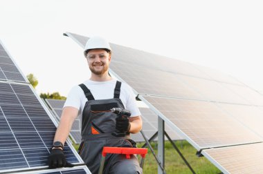Gülümseyen adam güneş panelleri kuruyor, elinde matkap var. Temiz enerji, yenilenebilir enerji ve sürdürülebilir kalkınma sunuluyor