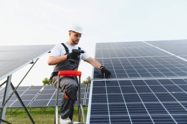 Merdivendeki erkek teknisyen, güneş çiftliğine fotovoltaik paneller yerleştirmek için matkap kullanıyor, güvenlik donanımı ve iş üniforması giyiyor.