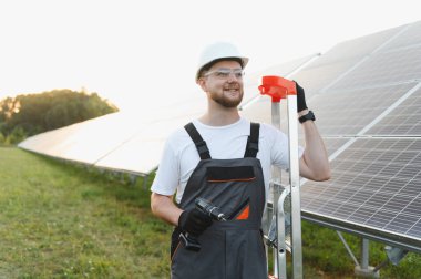 Güvenlik teçhizatı takan erkek teknisyen, elinde matkap, ve dışarıda güneş panellerinin yanında duran bir merdiven.