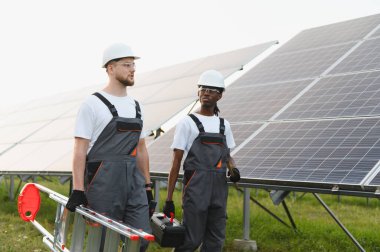 Çeşitli mühendisler alet ve merdiven taşıyor, güneş panellerinin yanında yürüyor, yenilenebilir enerji ve takım çalışmasını sembolize ediyor