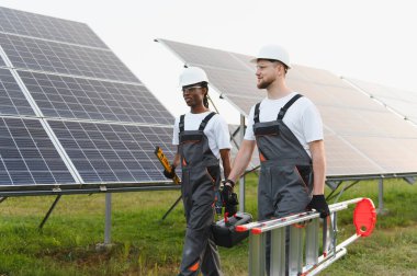 İki farklı mühendis, yeşil bir enerji çiftliğinde güneş panelleri kurmaya ya da muhafaza etmeye hazırlanan araçlar ve bir merdiven taşıyor.