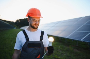 Başlık ve güvenlik gözlüğü takan mühendis, arka planda temiz enerji üreten güneş panelleri ile parlayan bir ampul tutuyor.