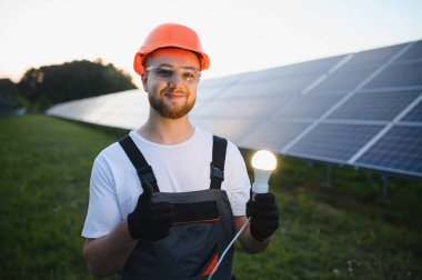 Genç teknisyen, başlık ve güvenlik gözlüğü takıyor, elinde ampul tutuyor ve güneş çiftliğinde baş parmak işareti yapıyor.