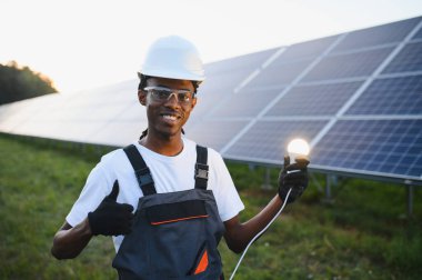 Gülümseyen adam yeşil, sürdürülebilir enerji için yeni bir fikir buluyor, güneş panellerinin yanında parlayan bir ampul tutuyor.