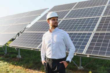 Güneş panellerinin önünde duran, sürdürülebilir enerji üretimi üzerinde çalışan, kasklı bir adam.